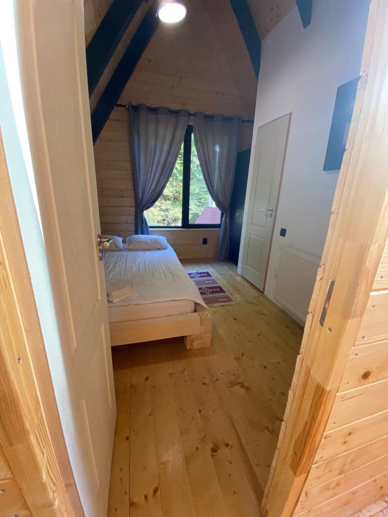 Dormitor cu peretele frontonului vopsit, fereastră spre pădure și pat matrimonial din lemn.