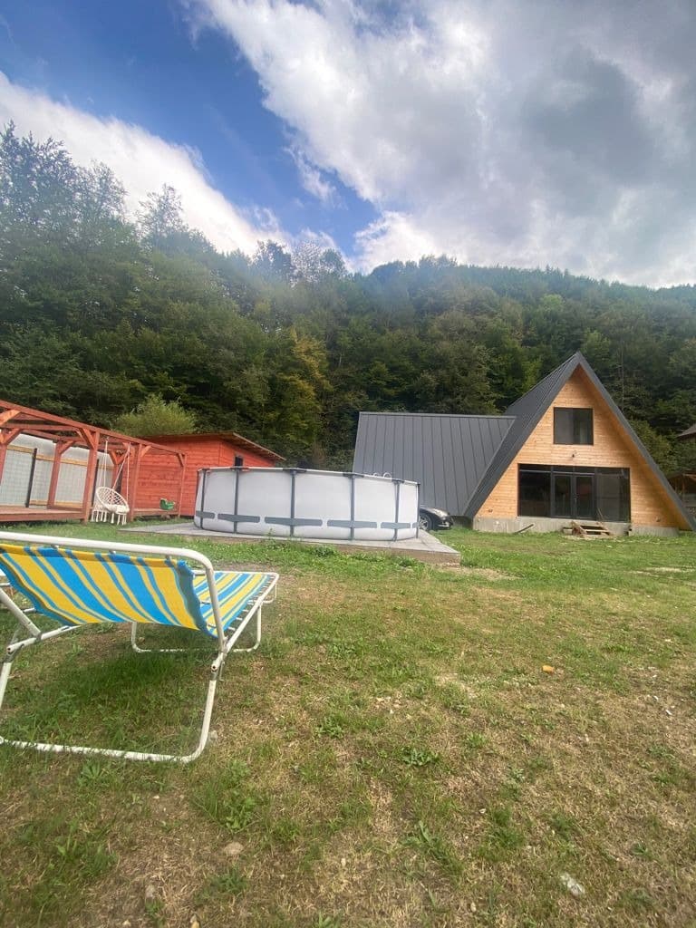 Cabana A-frame pe gazon, cu piscina alături și versantul împădurit în spate; în prim-plan un șezlong cu dungi albastre și galbene.