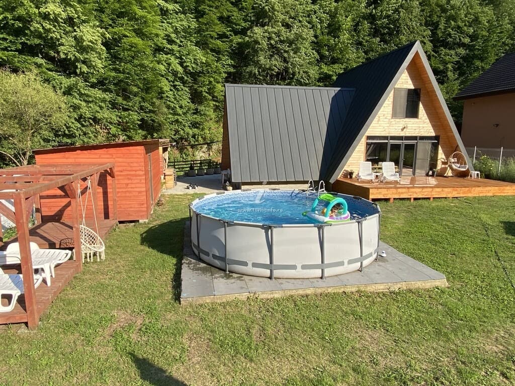 Cabana A-frame văzută din curte: acoperiș metalic grafit care coboară până la sol, fronton din lemn deschis cu glasvand, terasă din lemn și piscina rotundă pe platformă.