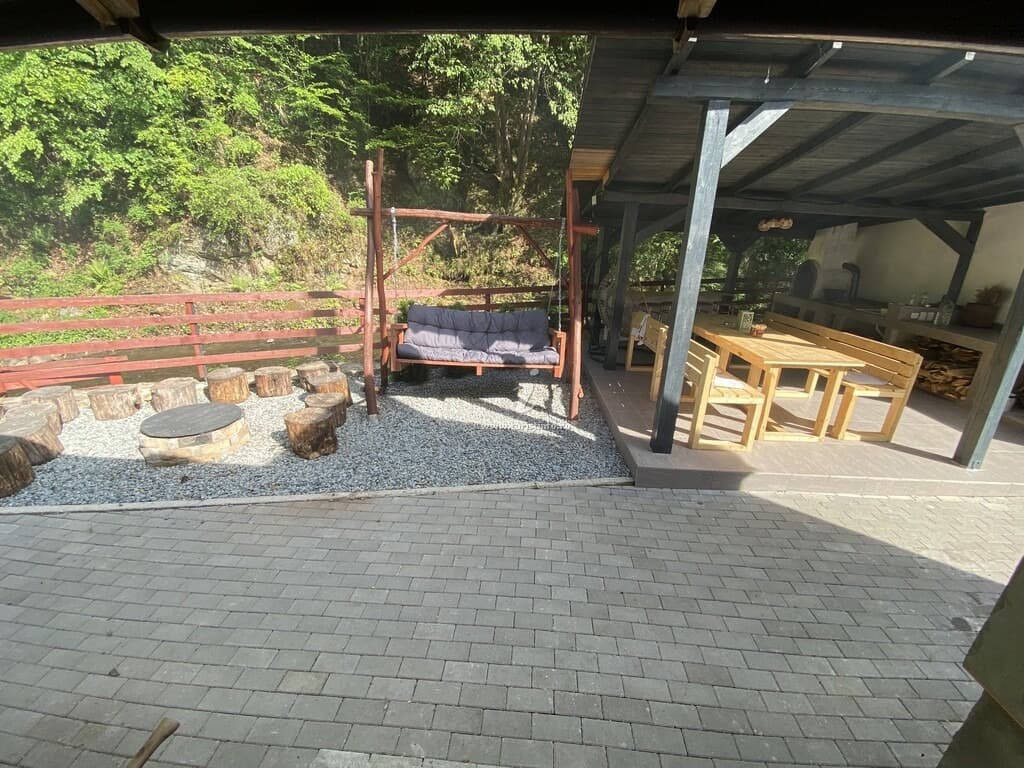 Zona acoperită din curte, cu masă lungă de lemn, bănci, vatră de foc înconjurată de scaune din bușteni și pietriș.