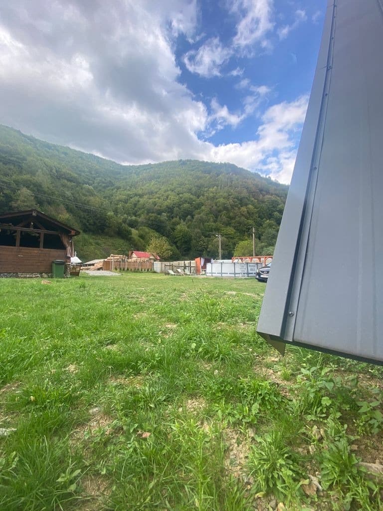 Acoperișul cabanei văzut de pe pajiște, cu dealurile împădurite ale văii în fundal.