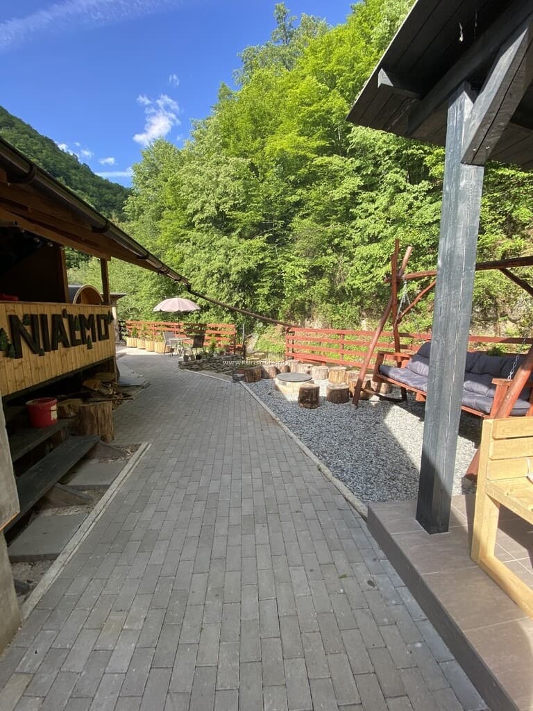 Aleea pavată care traversează curtea, cu literele verzi NIALMO și brăduții montați pe peretele de lemn din stânga, banchetă-balansoar și vatra de foc în dreapta.