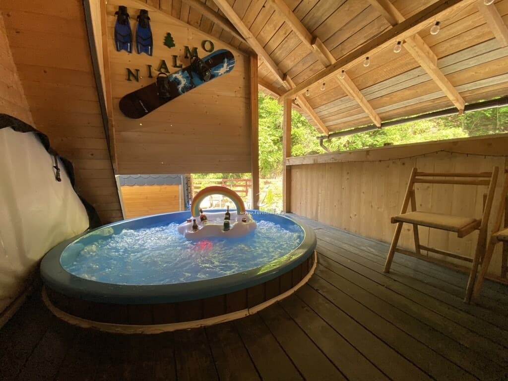 Jacuzzi rotund cu apă în mișcare, sub pavilionul acoperit din lemn; pe fronton, literele verzi NIALMO cu un brăduț, alături de un snowboard și labe de înot.