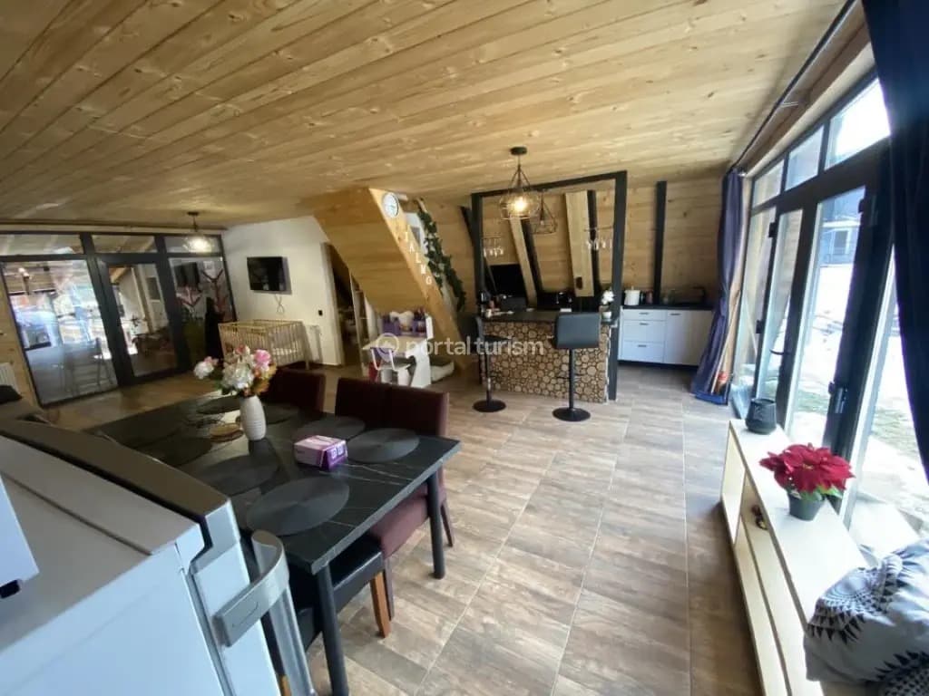 Livingul open-space pe toată lungimea cabanei: grinzi negre care traversează tavanul de lemn, masă de dining, perete vitrat spre vale și bucătăria în capăt.