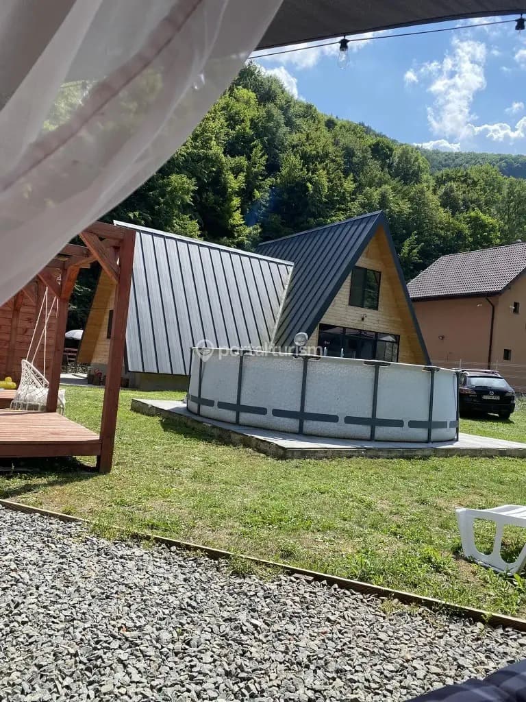 Piscina și cabana văzute de sub un baldachin cu perdele albe, cu versantul împădurit în fundal.