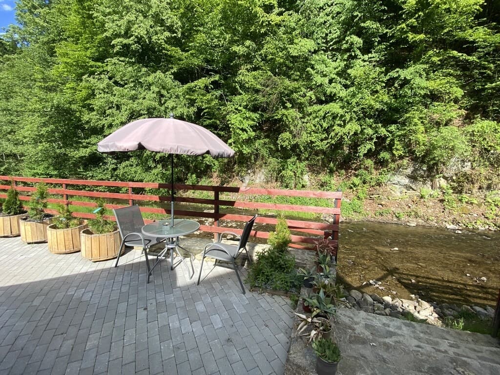 Terasă pavată deasupra apei, cu masă, scaune și umbrelă, mărginită de balustrada de lemn vopsită în cărămiziu.